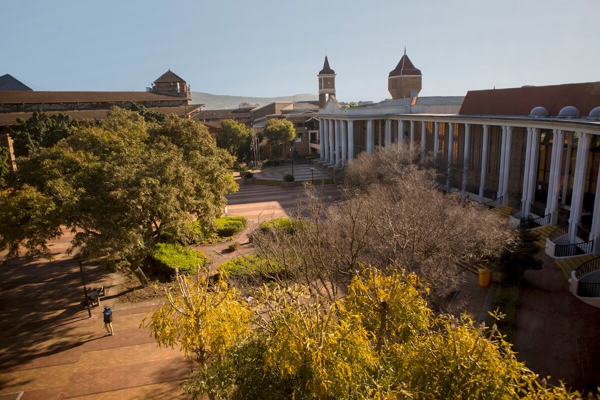 High-angle view of UWC's central piazza with brick paving, large leafy trees, and the long colonnaded Jakes Gerwel Hall curving along the right side.