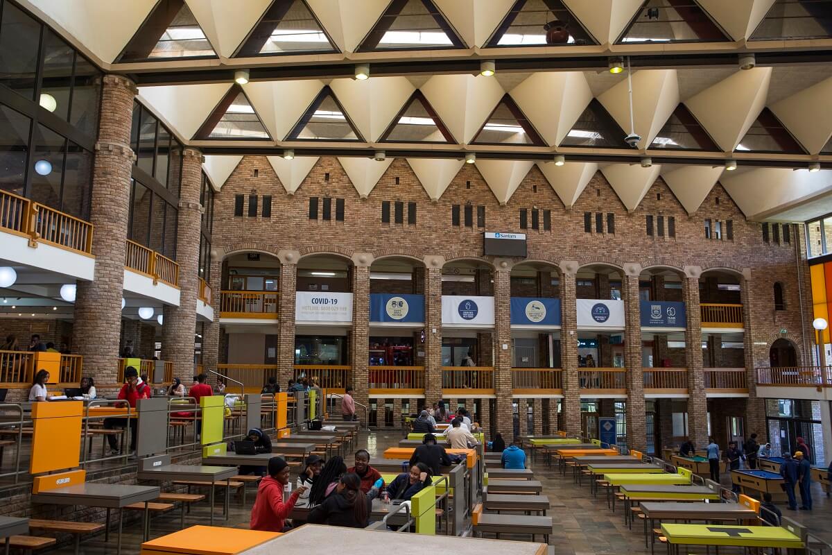 Interior of UWC Student Centre with brick pillars, high triangular ceiling, colorful orange and lime tables and benches, and students sitting, eating, and socializing.