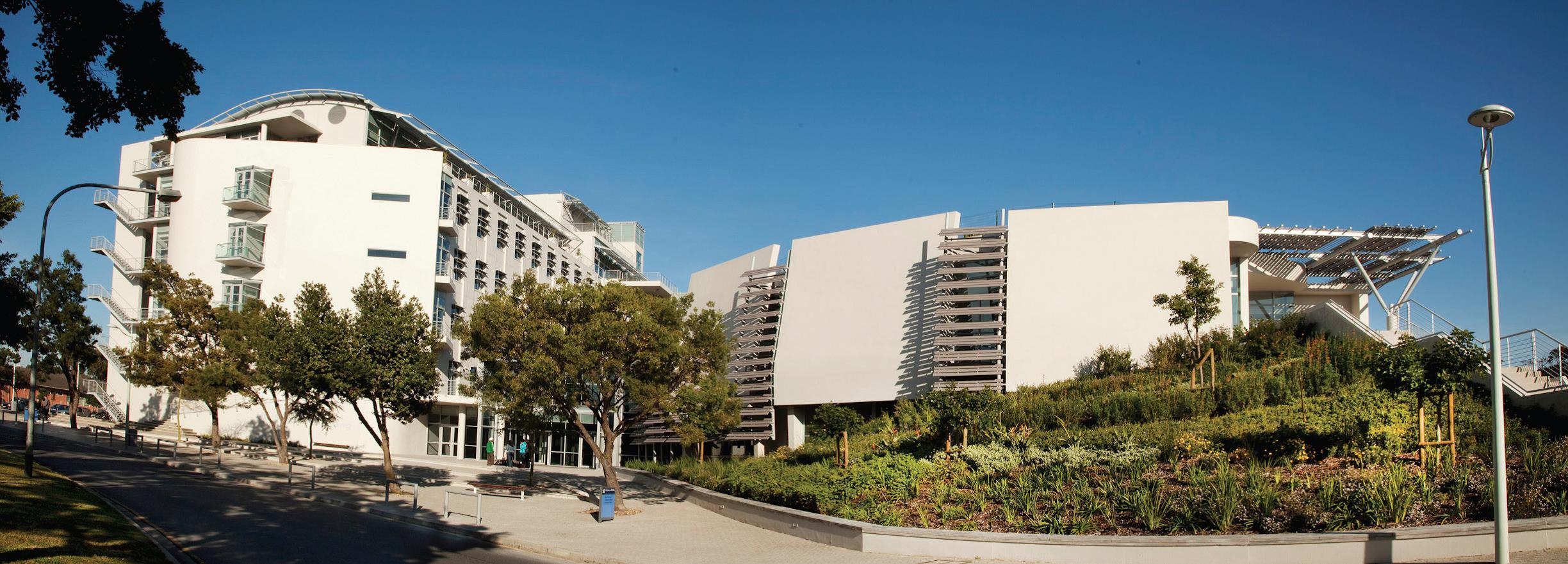 Modern white Life Sciences Building at UWC with curved architecture, glass balconies, and exterior staircases, surrounded by landscaped gardens and trees under a clear blue sky