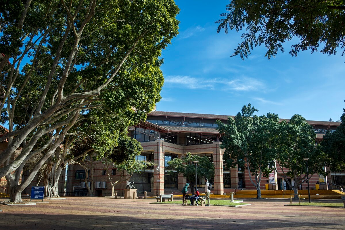 UWC Main Library, a two-storey brick building with large glass windows and striped pillars, shaded by large fig trees with students gathered on a bench in the forecourt.