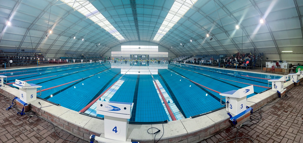Indoor Olympic-size swimming pool with bright blue water, lane dividers, and starting blocks numbered 2 to 5 under a large white arched tent roof with skylights.